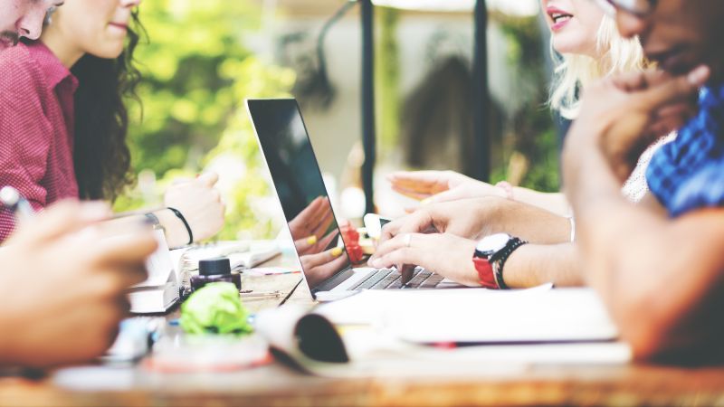 Zwei Personen mit einem Laptop sitzen weiteren Personen gegenüber