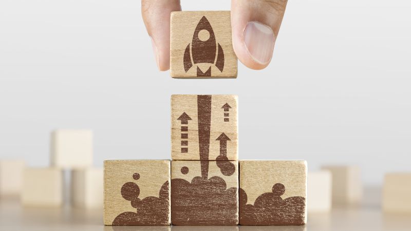 Zwei Finger heben einen mit kleiner Rakete bemalten Würfel