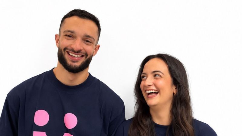 Hamdi Krichi und Anna-Luisa Raaf tragen jeweils ein T-Shirt mit pinkem Firmenlogo und lachen in die Kamera