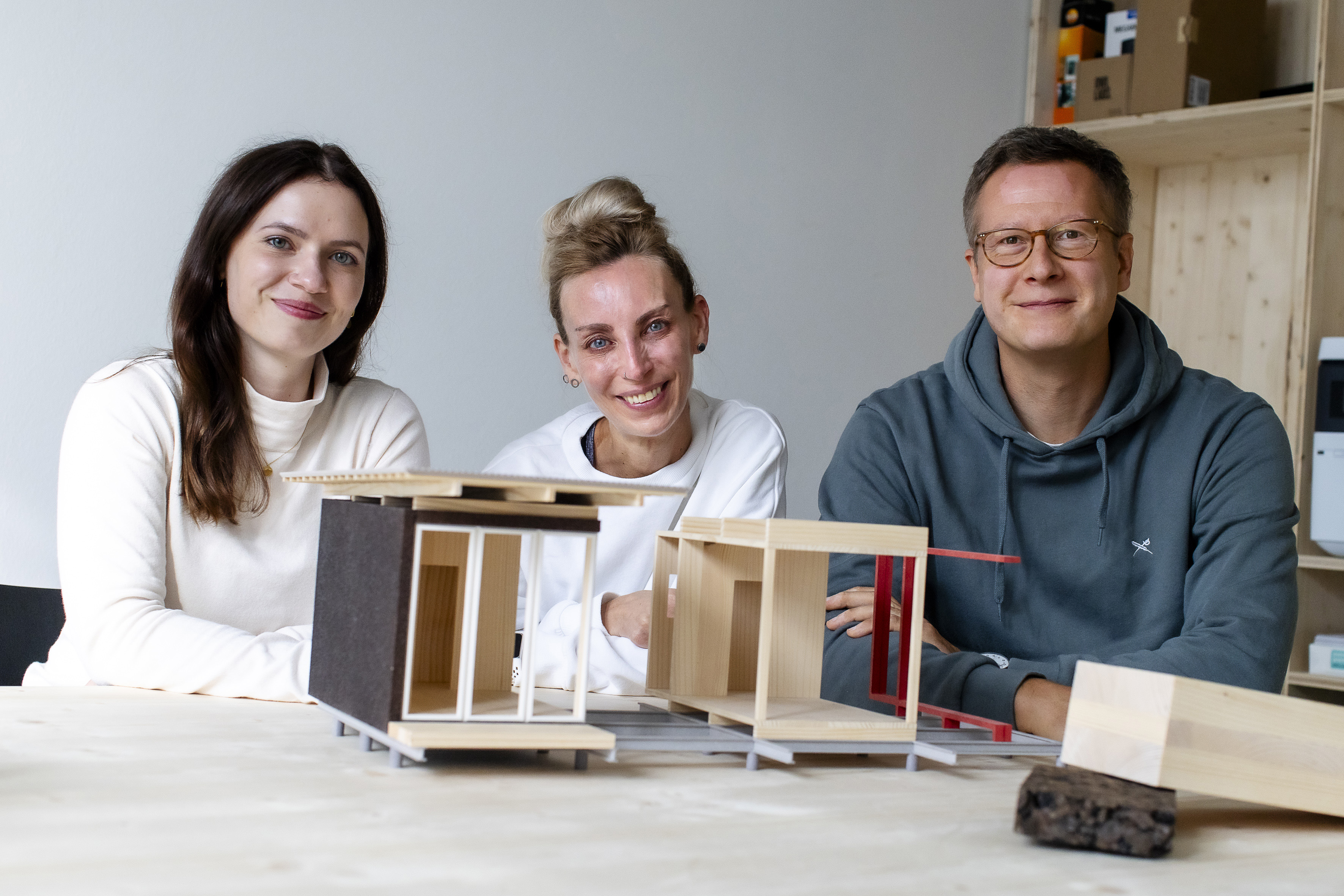 Die beiden Gründerinnen und ein Gründer sitzen vor einem kleinen Modell eines Modulhauses und lächeln in die Kamera