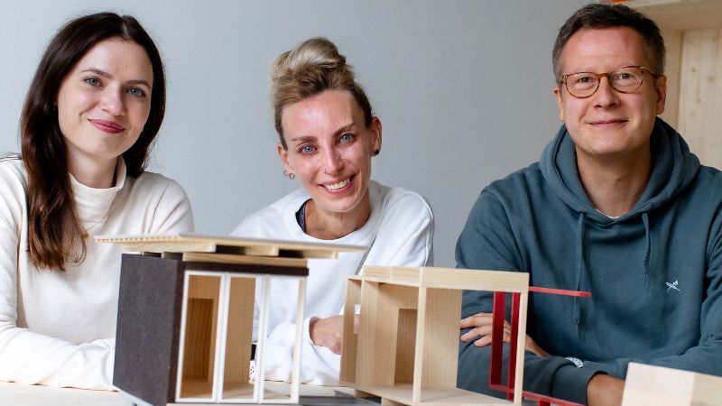 Die beiden Gründerinnen und ein Gründer sitzen vor einem kleinen Modell eines Modulhauses und lächeln in die Kamera