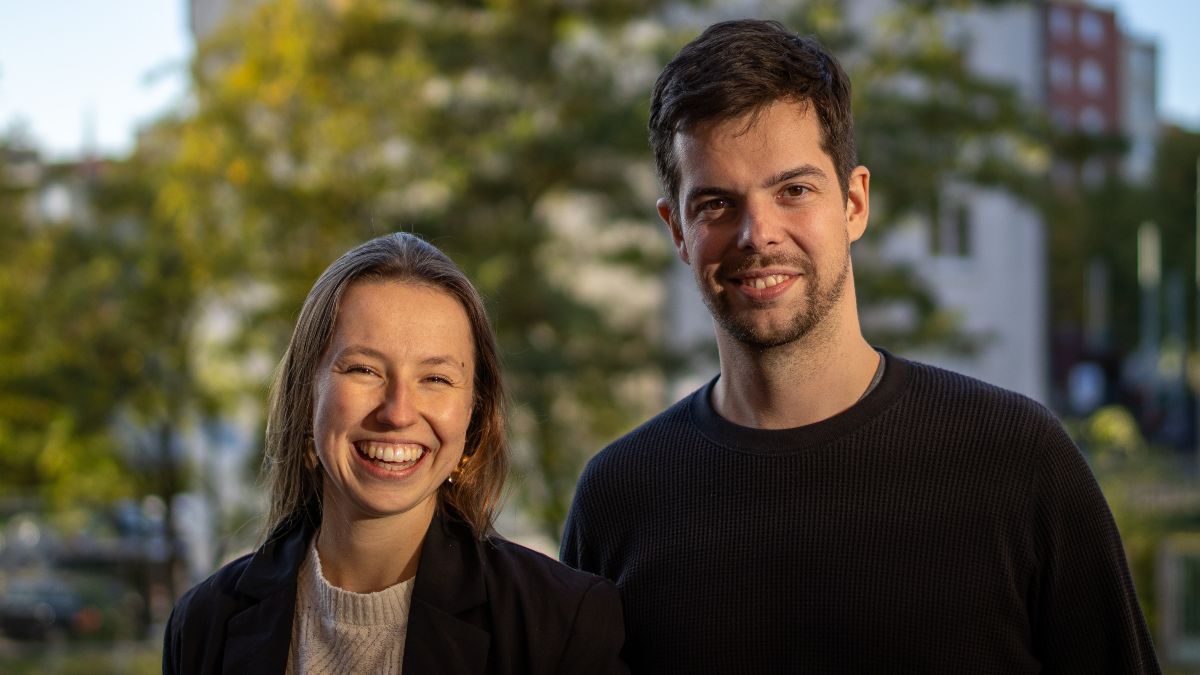 Dana Aleff und Erik Müller stehen nebeneinander draußen vor einem Baum und lächeln in die Kamera