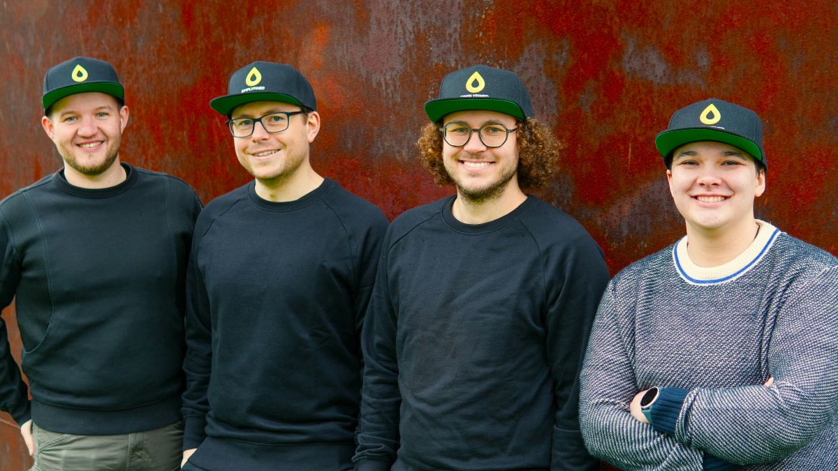 Die vier Gründer stehen nebeneinander vor einer rotbraunen Wand und lächeln in die Kamera