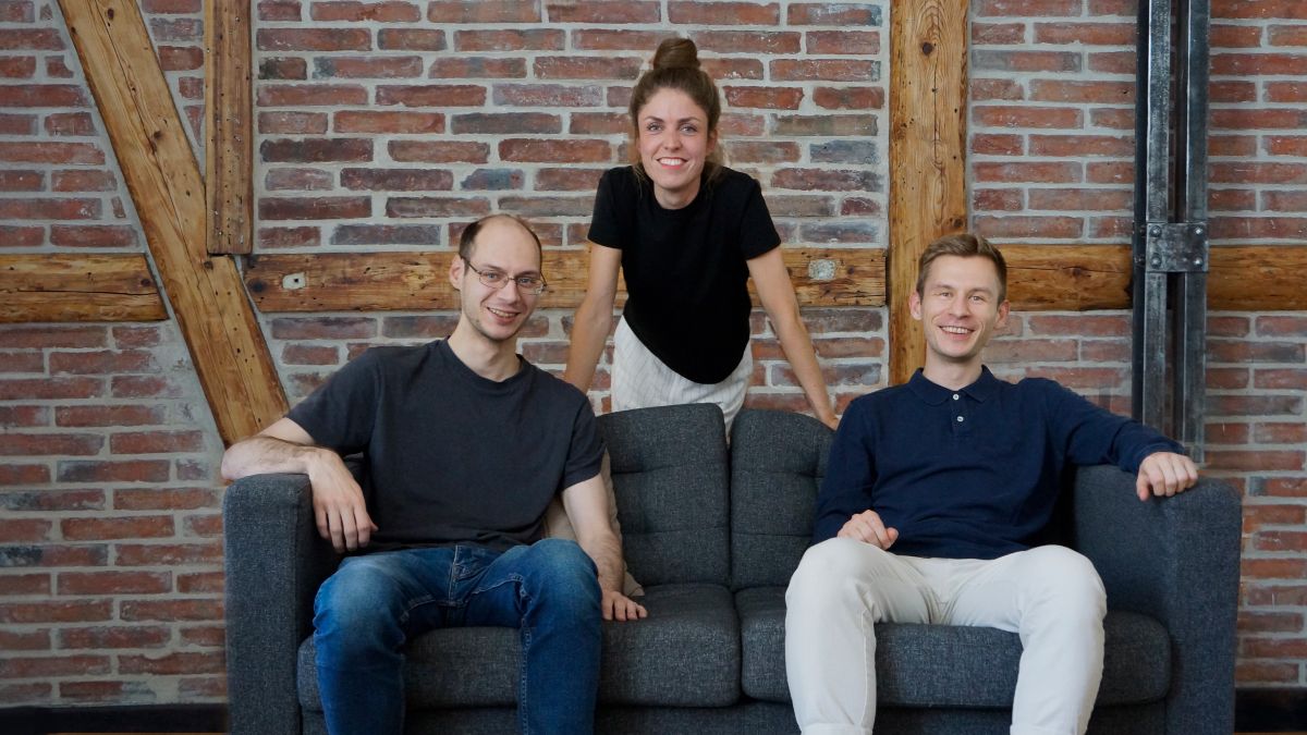 Die beiden Gründer sitzen auf einem Sofa und lächeln in die Kamera. Dahinter, in der Mitte, steht die Gründerin Sina Volkmann und lächelt ebenfalls in die Kamera.