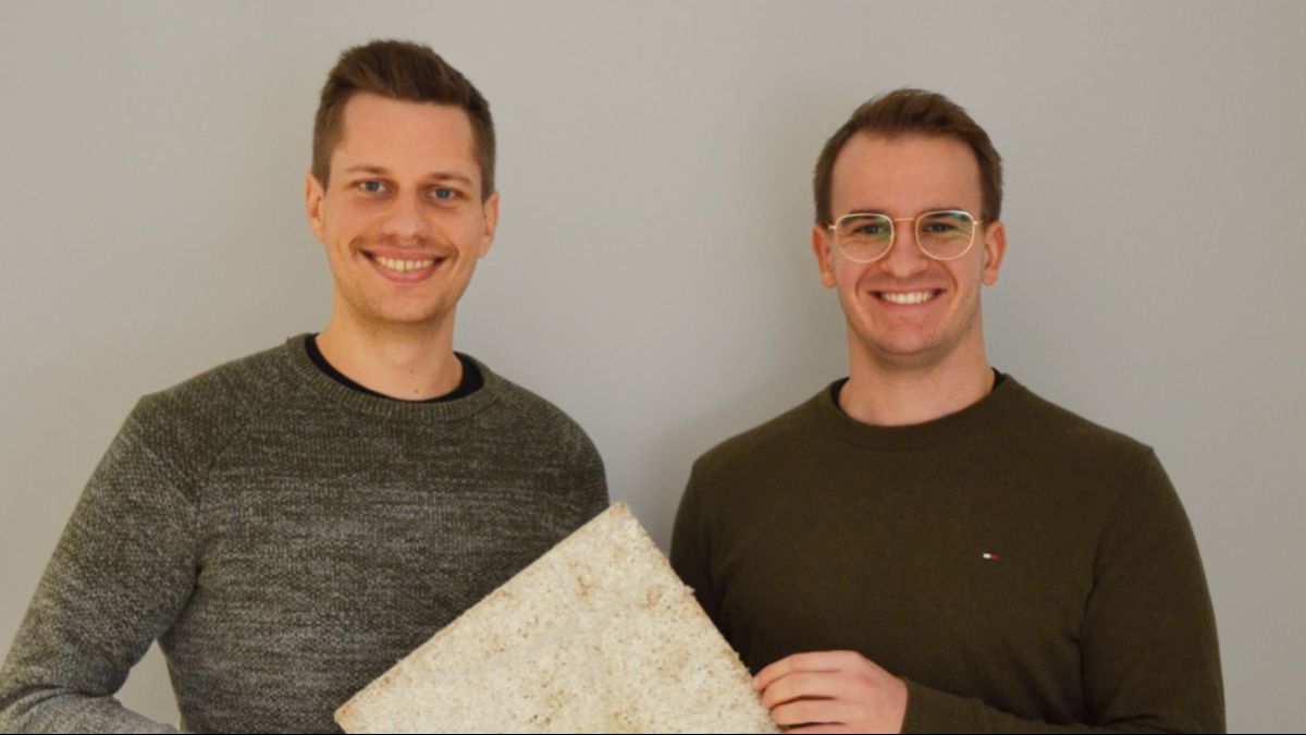 Zwei junge Männer stehen vor einer weißen Wand und lächeln in die Kamera.