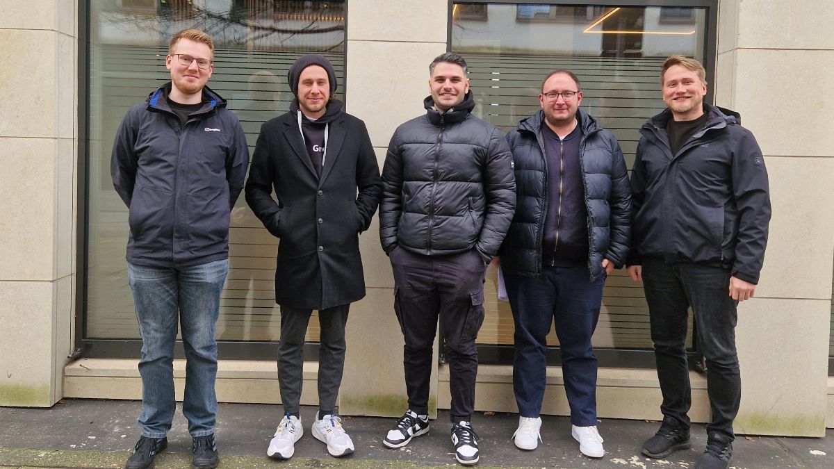 Fünf Männer im Alter von circa 25 bis 35 Jahren, alle in schwarz gekleidet, stehen draußen vor einem Fenster und lächeln in die Kamera.