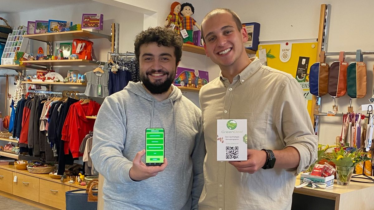 Die beiden jungen Gründer stehen in einem Bekleidungsgeschäft, blicken lächelnd in die Kamera und halten jeweils ein Smartphone mit Ihrer App in die Kamera